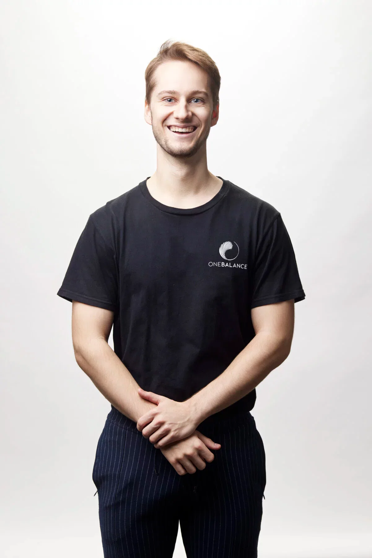 Lächelnder Mann im schwarzen T-Shirt steht vor weißem Hintergrund und hält die Hände locker vor dem Körper.