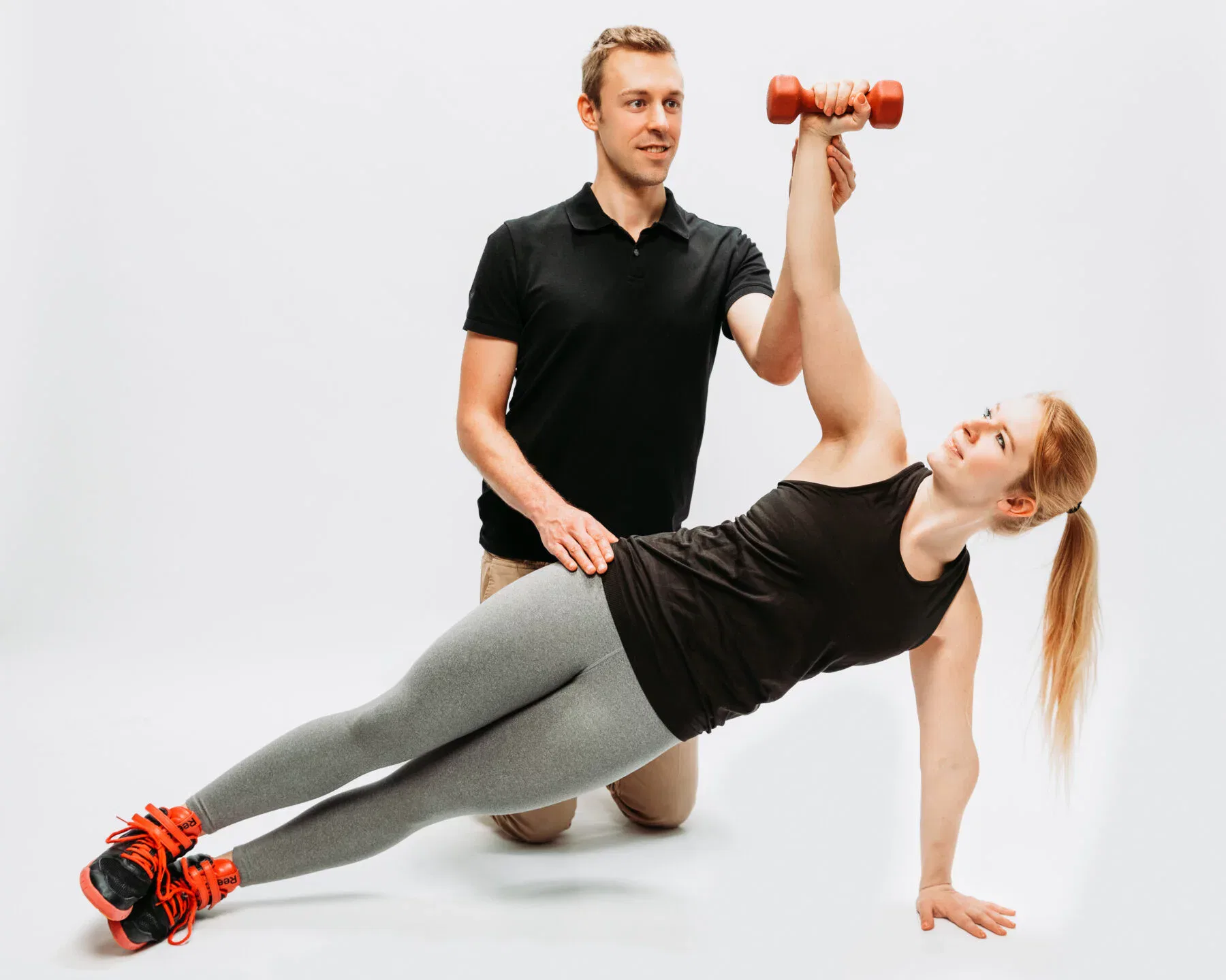 Personal Trainer betreut Frau beim Side-Plank und Schulterdrücken mit roter Kurzhantel im Fitnessstudio.