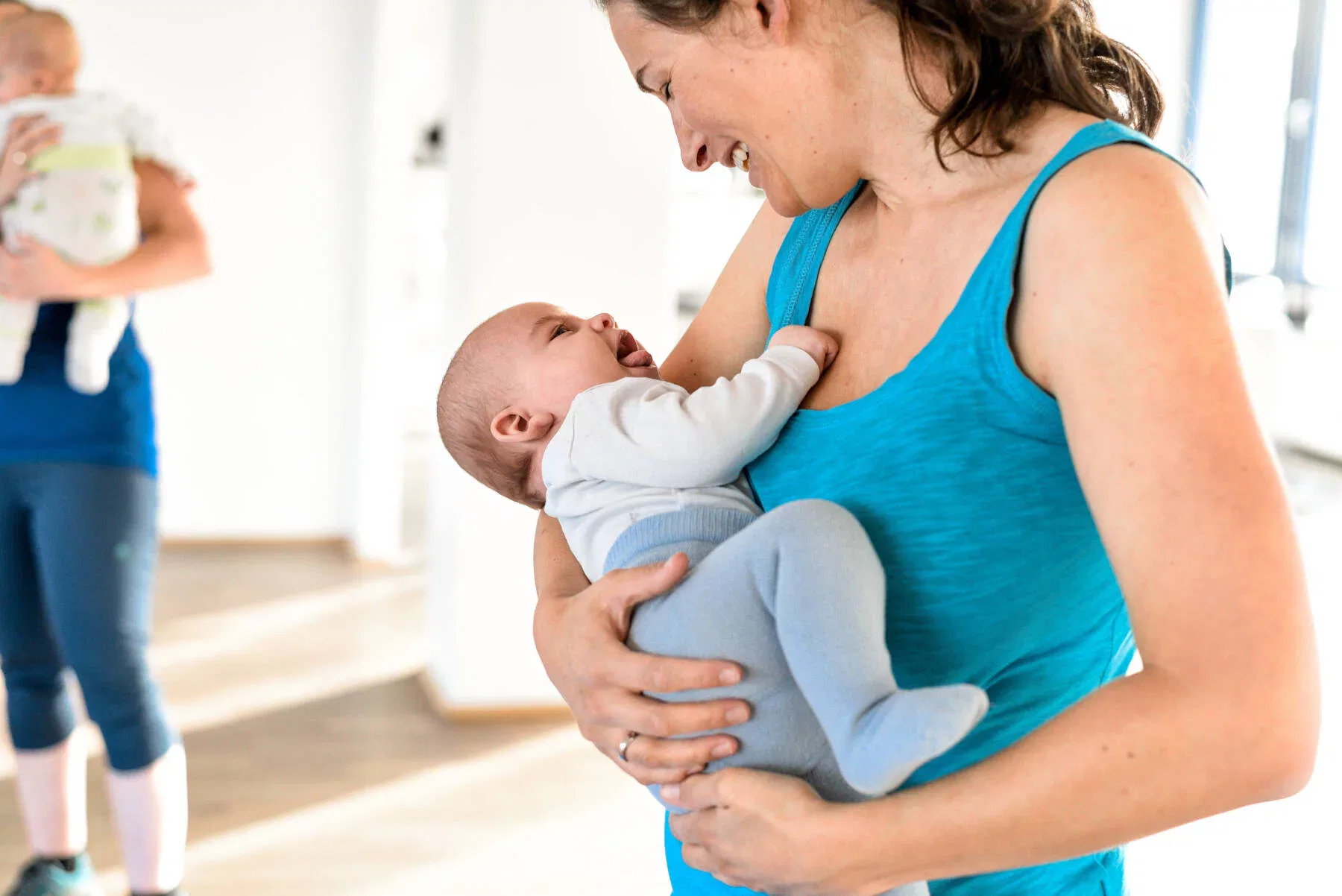 Eine Mutter hält ihr lachendes Baby im Arm und schaut es liebevoll an, im Hintergrund steht eine weitere Person mit Kind.