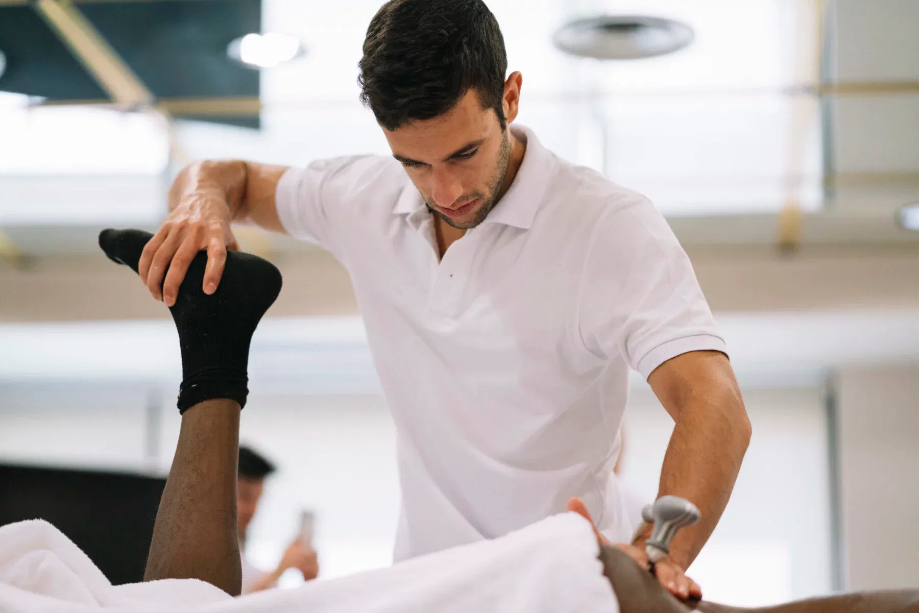 Physiotherapeut mobilisiert das Bein eines liegenden Patienten während einer Behandlung in einer hellen Praxis.